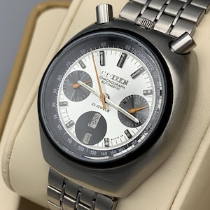 May include: A vintage Citizen Chronograph Automatic watch with a silver-tone stainless steel band. The watch face is black and silver with orange hands and a date display. The watch is displayed on a cream-colored stand.
