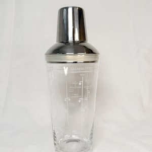 May include: A clear glass cocktail shaker with a stainless steel cap. The glass body features white printed text and measurement lines for a Manhattan cocktail, including labels for ingredients like red vermouth and Angostura bitters.