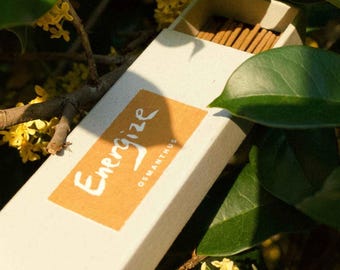 May include: A box of incense sticks with a label that reads "Energize Osmanthus". The box is white with a brown label and is sitting on a branch of a plant with yellow flowers.