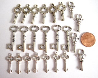 May include: A collection of 20 silver-toned key charms, featuring various ornate designs. The charms are approximately the size of a US penny.