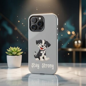 Könnte beinhalten: Hellgraue Handyhülle mit einem Cartoon-Hund mit schwarz-weißem Fell, rotem Halsband und blauen Augen. Der Text "Stay Strong" ist darunter gedruckt. Die Hülle hat ein Kameramodul mit drei Linsen.