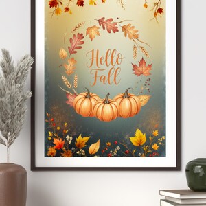 Könnte beinhalten: Ein gerahmter Kunstdruck mit Herbstthema. Das Kunstwerk zeigt einen Kranz aus Herbstblättern und drei orangefarbenen Kürbissen. Die Worte "Hello Fall" sind in eleganter Schrift geschrieben. Die Farbpalette umfasst Orange-, Gelb- und Brauntöne.