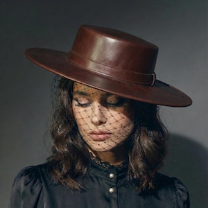 May include: A brown leather hat with a wide brim and a dark veil. The hat has a cylindrical crown and a band around the base. The veil is a fine black mesh. The hat is worn over dark hair and a black blouse.