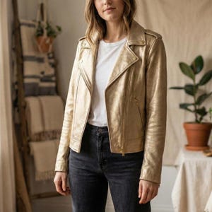 Puede incluir: Chaqueta de cuero dorada con cremallera asimétrica. La chaqueta presenta un cuello con solapas, charreteras y una silueta entallada. La modelo lleva una camiseta blanca y vaqueros oscuros.