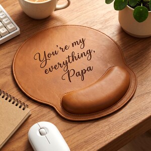 Puede incluir: Una alfombrilla de ratón redonda de piel sintética color canela con un reposamuñecas integrado. La superficie presenta el texto negro en cursiva "You're my everything, Papa" impreso en el centro. Tiene un acabado mate liso y bordes cosidos.