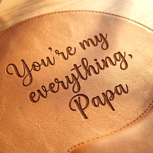 Könnte beinhalten: Ein runder, hellbrauner Ledergegenstand mit der eingravierten Aufschrift "You're my everything, Papa" in einer dunklen Schreibschrift. Die Oberfläche hat eine sichtbare Narbung und ist entlang der Kante mit passendem Garn vernäht.