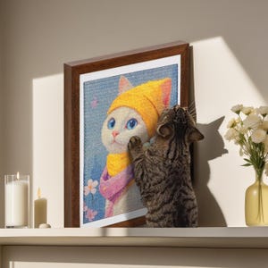 Pode incluir: Uma obra de arte emoldurada com uma ilustração de um gato com chapéu e cachecol amarelos. Um gato tigrado real interage com a imagem emoldurada. A obra de arte tem uma moldura de madeira castanha e é exibida numa prateleira branca com velas e um vaso de flores.