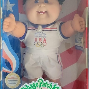 Può includere: Bambola Cabbage Patch Kids OlympiKids Special Edition nella confezione originale. La bambola indossa un'uniforme sportiva bianca e blu con il logo olimpico USA. La scatola presenta accenti patriottici rossi, bianchi e blu.
