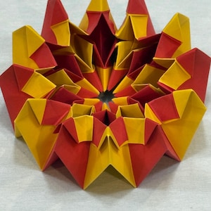 May include: A vibrant, geometric origami sculpture in red and yellow. The paper art piece is a symmetrical, star-like shape with multiple layers and folds. The colors create a striking contrast, highlighting the intricate design.