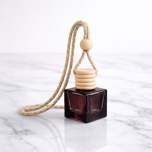 May include: A dark purple, square-shaped car air freshener with a wooden cap and a beige rope for hanging. The wooden cap has a layered design. The air freshener is displayed on a white marble surface.