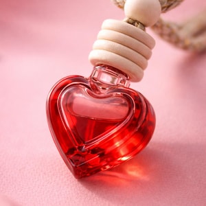 May include: A heart-shaped, red glass bottle filled with a red liquid, suspended by a beige rope and wooden accents. The bottle is a decorative air freshener, perfect for a car or small space. The background is a soft pink.