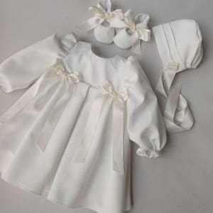 May include: A white baby dress set featuring a long-sleeved dress with ribbon bows, matching booties with bows, and a bonnet with ribbon ties. The set is made of a soft, light-colored fabric.
