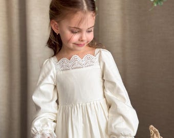 Könnte beinhalten: Ein junges Mädchen trägt ein weißes Kleid mit Spitzenbesatz und Puffärmeln. Sie hält eine Gießkanne in der Hand und schaut nach unten. Das Kleid hat einen Stufenrock und einen quadratischen Ausschnitt. Das Mädchen steht vor einem braunen Hintergrund mit Grünpflanzen.