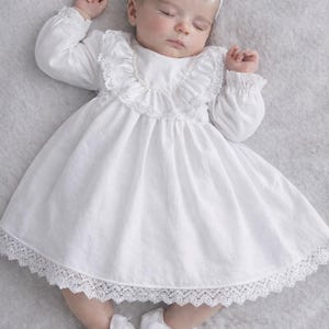 May include: A sleeping baby wearing a white dress with lace trim, long sleeves, and a ruffled collar. The baby also wears a white bow headband and matching shoes with bows. The baby is lying on a gray surface.
