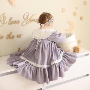 Può includere: Una bambina che indossa un vestito lavanda con bordino in pizzo bianco e un colletto in pizzo bianco. Indossa una corona di fiori ed è seduta su una cassettiera bianca.