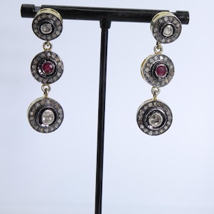 May include: A pair of silver and gold dangle earrings with three round stones. The top stone is clear, the middle stone is red, and the bottom stone is clear. The stones are surrounded by smaller clear stones.