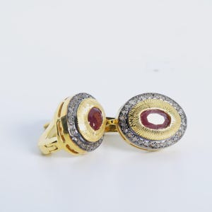 May include: A pair of gold earrings with oval-shaped red gemstones surrounded by a halo of smaller diamonds.