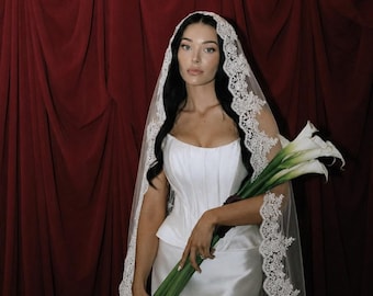 May include: A white satin corset-style dress with a structured bodice and a floor-length skirt, paired with a long, sheer veil featuring intricate white lace trim along the edges. The ensemble is accessorized with a bouquet of white calla lilies.