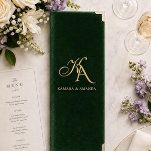 May include: A rectangular dark green velvet menu cover featuring gold foil lettering with the monogram KA and the names Kamara & Amanda. The cover has silver-toned metal corner protectors and is positioned next to a printed paper menu card.