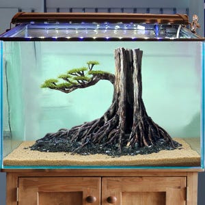 Può includere: Un acquario con un elemento centrale a forma di albero artificiale dettagliato. L'albero presenta un tronco marrone strutturato e radici esposte, con una chioma verde e fogliosa. L'acquario ha un'illuminazione e poggia su un mobile in legno.