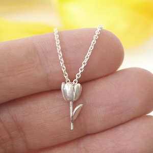 May include: A silver tulip pendant necklace. The pendant is a detailed miniature of a tulip flower with a stem and leaf. The necklace is displayed against a blurred yellow and white background.