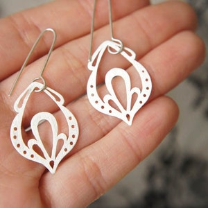 以下が含まれることがあります： 繊細で複雑なデザインのシルバー製のイヤリングのペア。イヤリングは、花柄の涙の形をしています。