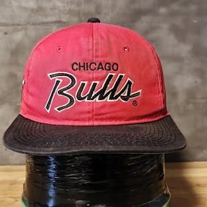 May include: A red baseball cap with a black brim featuring the embroidered text CHICAGO Bulls in black and white lettering on the front. The cap has a structured crown with a small black button at the top and ventilation eyelets on the panels.