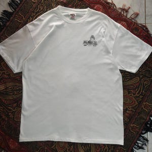 Puede incluir: Una camiseta blanca de manga corta con un pequeño gráfico en la parte superior derecha del pecho. El gráfico incluye una pequeña ilustración y el texto Microsoft SQL Server 7.0. La camiseta tiene cuello redondo y un diseño estándar.