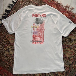 May include: A white short-sleeve t-shirt featuring a graphic on the back with the red text MET-Rx and the words High Performance Series. The design includes a faint image of a muscular figure and various text elements like HMB, Ultra Myosyn, and MKP.