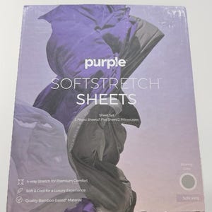 Op de afbeelding: Een doos met paarse Softstretch lakens. De verpakking heeft een paars en grijs abstract ontwerp. De tekst op de doos luidt "Softstretch Sheets" en bevat details over de lakenset, waaronder twee hoeslakens, een plat laken en twee kussenslopen. De kleur is "Stormy Grey".