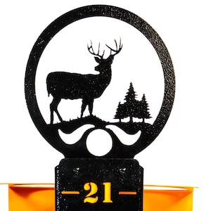 May include: A black metal deer silhouette with trees in a circle on top of a black metal ruler with orange numbers from 13 to 21. The ruler has two white and yellow arrows.