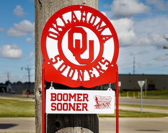 Könnte beinhalten: Rotes Metallschild mit den Worten "OKLAHOMA SOONERS" in einem kreisförmigen Design und einem kleineren Schild darunter mit "BOOMER SOONER". Das Schild ist auf einem roten Metallrahmen in einer Rasenfläche montiert.