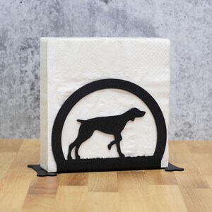 May include: Black metal napkin holder with a silhouette of a dog in a circle. The holder is on a wooden surface.