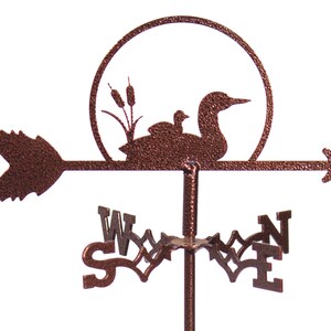 May include: A copper-colored metal wind vane featuring a loon and its chick perched on a branch with cattails. The wind vane has a circular design with a sun behind the loon and chick. The wind vane also has a directional arrow and compass points.