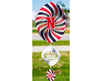 May include: A red, white, and black spiral wind spinner with a large "N" in the center and the words "Go Big Red" on a football-shaped spinner below. The bottom spinner is a smaller red, white, and black spiral with the word "Huskers" on it.