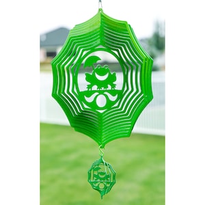 May include: Green metal wind spinner with a frog design. The spinner has multiple layers and is suspended from a hook. The bottom of the spinner has a smaller, round, green metal piece with a frog design.