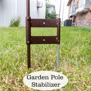 K&ouml;nnte beinhalten: Ein brauner Gartenmast-Stabilisator aus Metall mit zwei Metallpfosten. Der Stabilisator ist so konzipiert, dass er hilft, einen Gartenflaggenmast aufrecht zu halten.