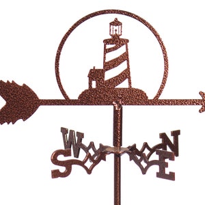 May include: A copper-colored metal weather vane featuring a lighthouse with a striped tower and a small house at its base. The lighthouse is set within a circular frame and the vane has a directional arrow with a feathered design. The compass directions are labeled with the letters N, S, E, and W.