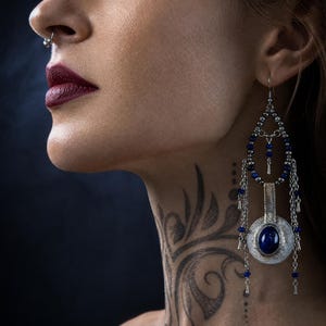 Puede incluir: Un pendiente largo colgante de plata con una piedra azul ovalada central en un marco circular ornamentado, acentuado con pequeñas cuentas azules, cadenas de plata y una sección superior en forma de lágrima con cuentas de plata.