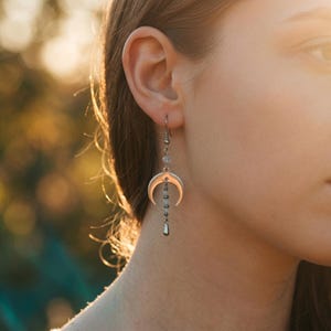 Puede incluir: Pendientes plateados con forma de media luna y un dije colgante en forma de lágrima. Los pendientes presentan una serie de pequeñas cuentas y un diseño delicado. Los pendientes se muestran en una oreja.