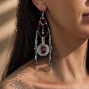 Puede incluir: Un pendiente largo y colgante con un marco en forma de lágrima de pequeñas cuentas rojas, un medallón circular plateado con una piedra roja ovalada y varias cadenas plateadas que cuelgan, decoradas con cuentas rojas y gotas plateadas.