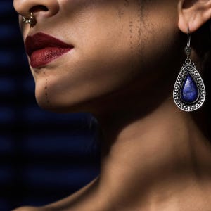 Puede incluir: Un pendiente colgante de plata con una piedra azul en forma de lágrima engastada en un marco de metal texturizado y ornamentado. El pendiente cuelga de un gancho sencillo y muestra intrincados detalles de metal alrededor de la piedra central.