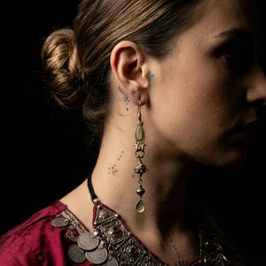 Puede incluir: Pendientes largos y ornamentados de latón, con un colgante en forma de lágrima. Los pendientes presentan detalles intrincados y están suspendidos de un gancho. La imagen muestra un primer plano de los pendientes sobre un fondo oscuro, sugiriendo un estilo vintage.