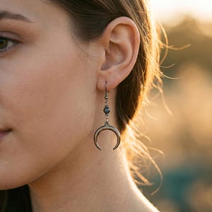 Puede incluir: Pendientes plateados con forma de media luna con un pequeño detalle de cuentas oscuras. Los pendientes cuelgan de un gancho simple y presentan un diseño pulido y curvado. Un accesorio de temática celestial.