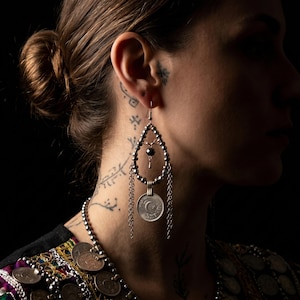 May include: Silver teardrop earrings with a beaded design and dangling chains. Each earring features a coin with a crescent moon and star. The earrings are worn with a colorful embroidered top.