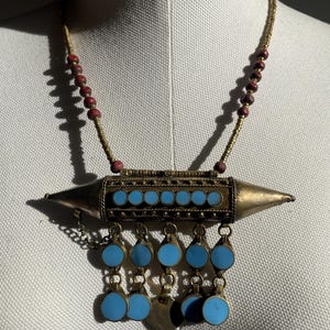 May include: A vintage necklace with a central silver-toned pendant featuring turquoise-colored circular inlays and dangling turquoise accents. The necklace has a beaded gold-colored chain with red beads interspersed throughout.