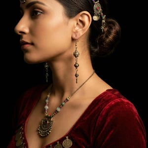 Puede incluir: Un conjunto de joyas ornamentado de estilo antiguo con pendientes largos colgantes, un collar de cuentas con un colgante y una diadema. Las joyas son de metal con un acabado deslustrado. La mujer lleva un top de terciopelo rojo.