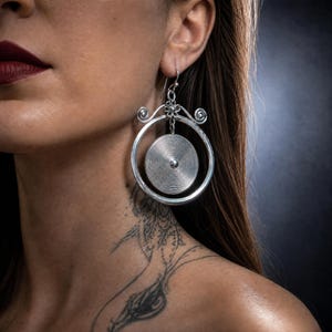 Puede incluir: Un pendiente colgante de tono plateado que presenta un gran aro exterior con volutas decorativas en la parte superior. Del centro del aro cuelga un disco circular más pequeño con un patrón de espiral concéntrico fino y un punto central.