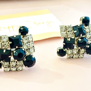 May include: A pair of square-shaped earrings with a silver-tone metal setting. Each earring features a cluster of clear, sparkling rhinestones and dark navy blue, faceted stones in various shapes. The earrings are displayed on a light pink surface.