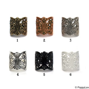 Peut inclure: Six bagues en métal ornées de différentes couleurs : laiton antique, cuivre, argent, noir et blanc. Chaque bague présente un motif floral filigrané détaillé. Les bagues sont présentées sur fond blanc.